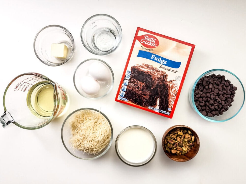 Top-down view of ingredients for baking: brownie mix, eggs, butter, oil, water, chocolate chips, milk, shredded phyllo dough, and chopped pistachios in separate bowls on a white surface.