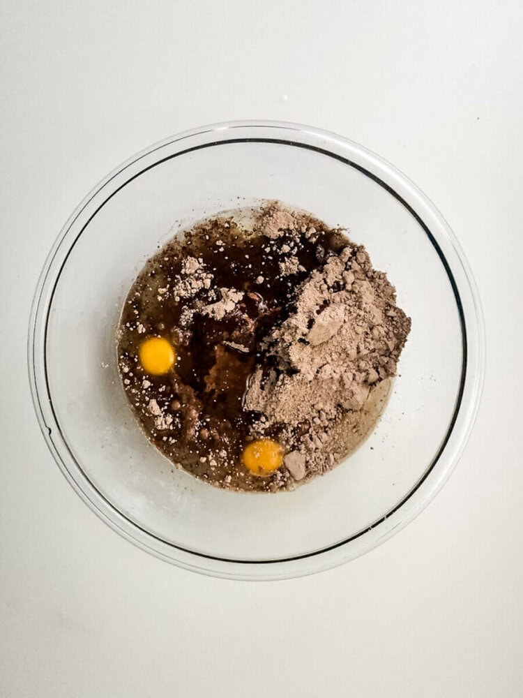 A glass bowl containing two eggs, cocoa powder, and liquid ingredients, ready to be mixed for baking.