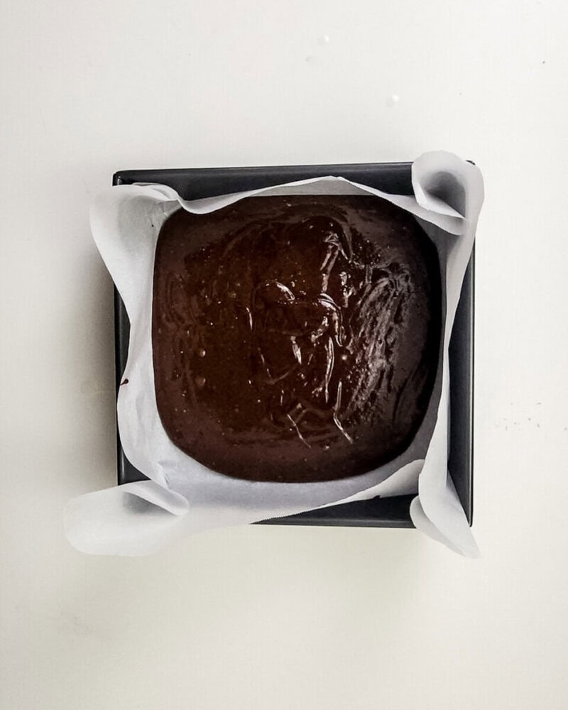 A square baking pan lined with parchment paper, filled with unbaked chocolate cake batter, sits on a white surface.