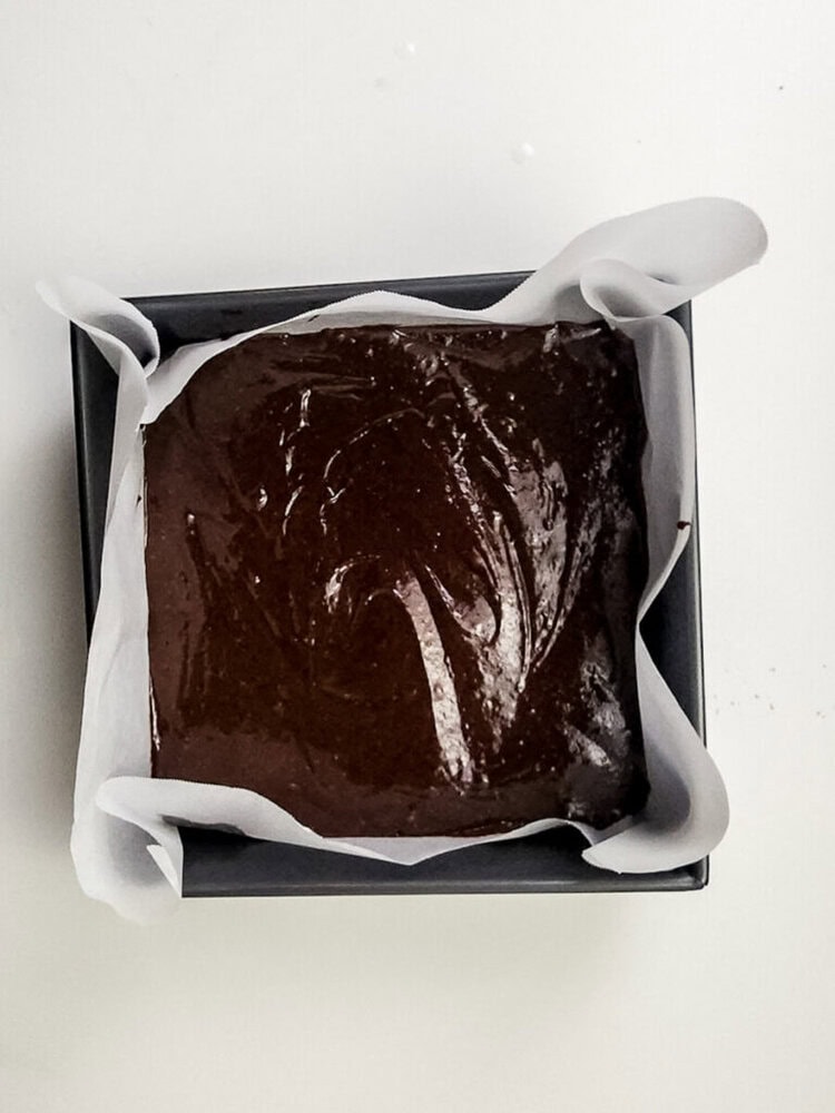 A square baking tin lined with parchment paper is filled with unbaked chocolate brownie batter.