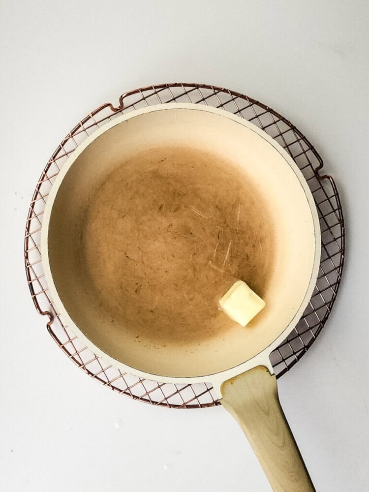A beige frying pan with a wooden handle and a piece of butter inside sits on a round wire cooling rack against a white background.