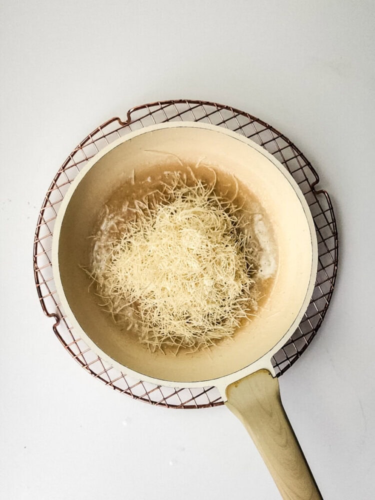 A beige pan with a wooden handle sits on a wire rack, containing melted butter and a pile of shredded cheese.