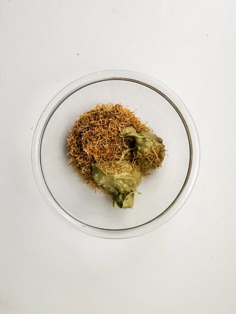 A glass bowl containing a pile of dried brown threads and two greenish dried flower buds, set on a plain white background.