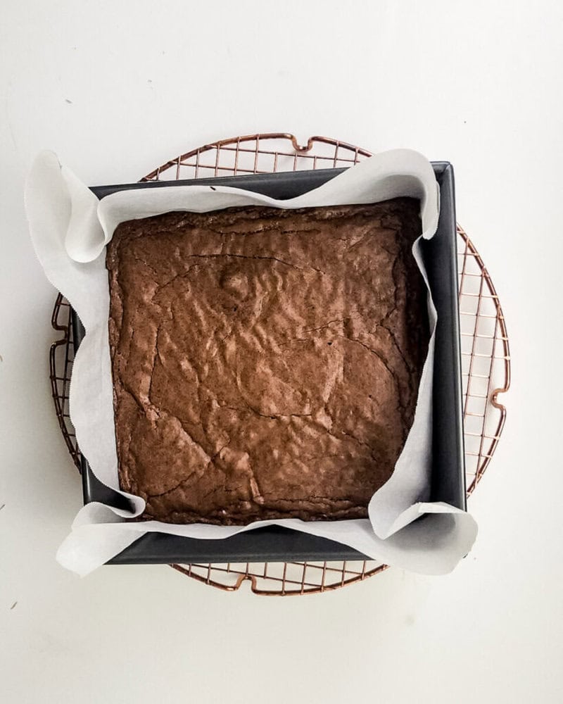 A square pan of baked brownies with parchment paper lining sits on a round cooling rack against a white background.