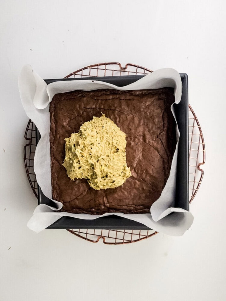 A square baking pan lined with parchment paper contains a baked brownie base with a mound of unspread yellowish frosting in the center, set on a cooling rack.