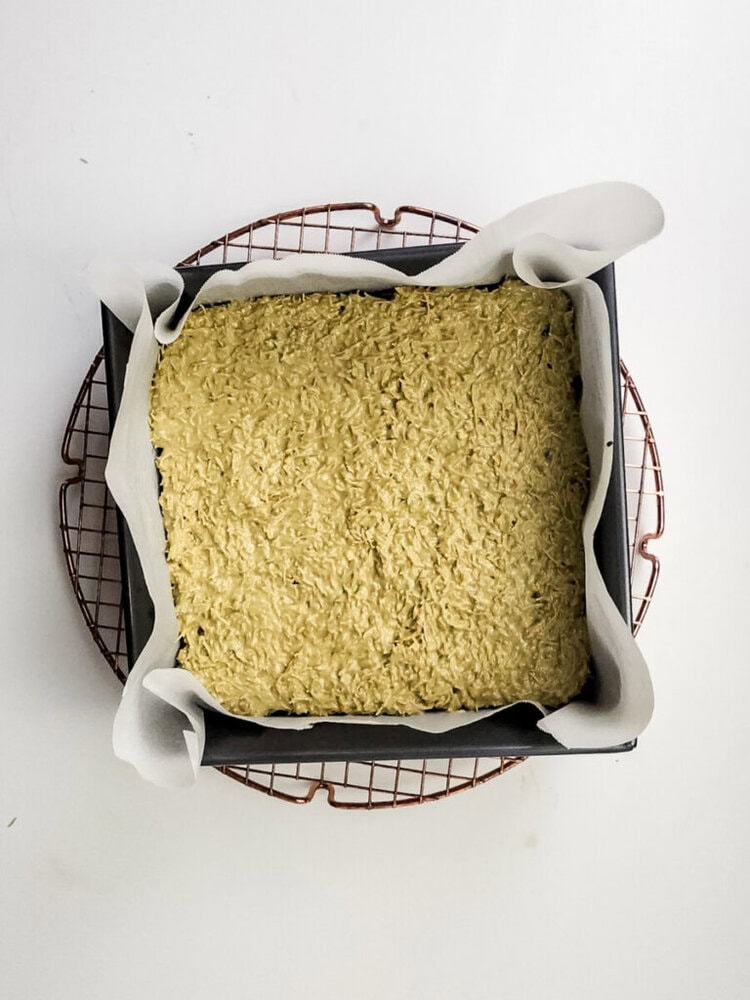 A square baking pan lined with parchment paper holds an unbaked oat mixture, resting on a round cooling rack against a white background.