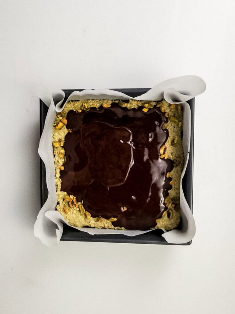 A square baking pan lined with parchment paper holds a partially baked oat crust topped with a layer of chocolate.