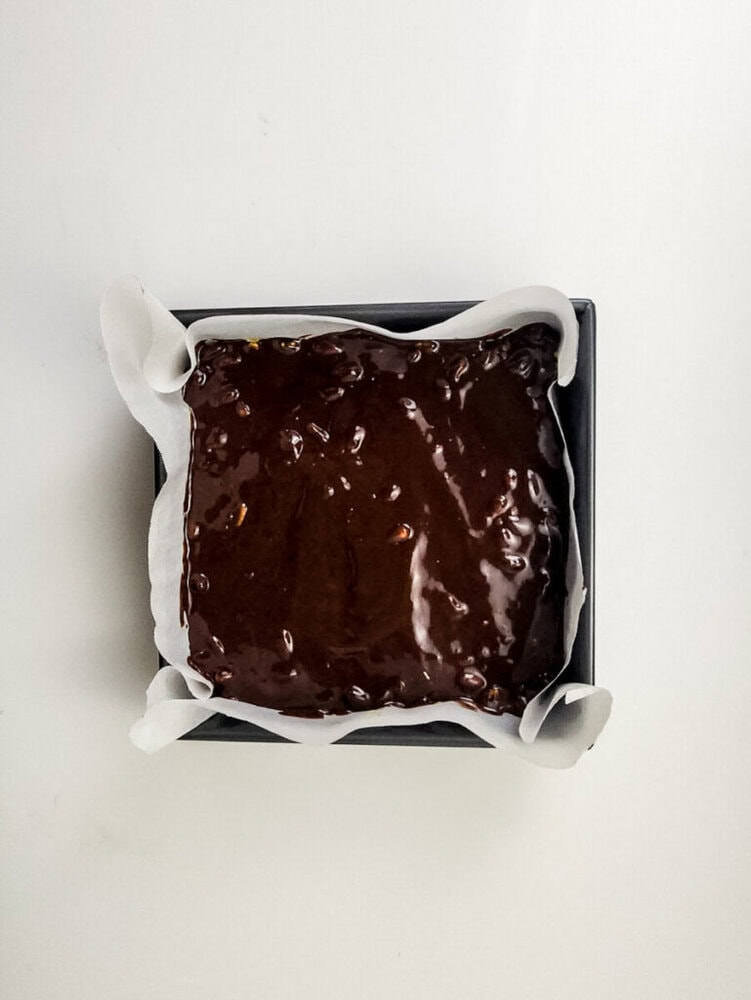 A square baking pan lined with parchment paper filled with unbaked chocolate brownie batter, viewed from above on a white surface.