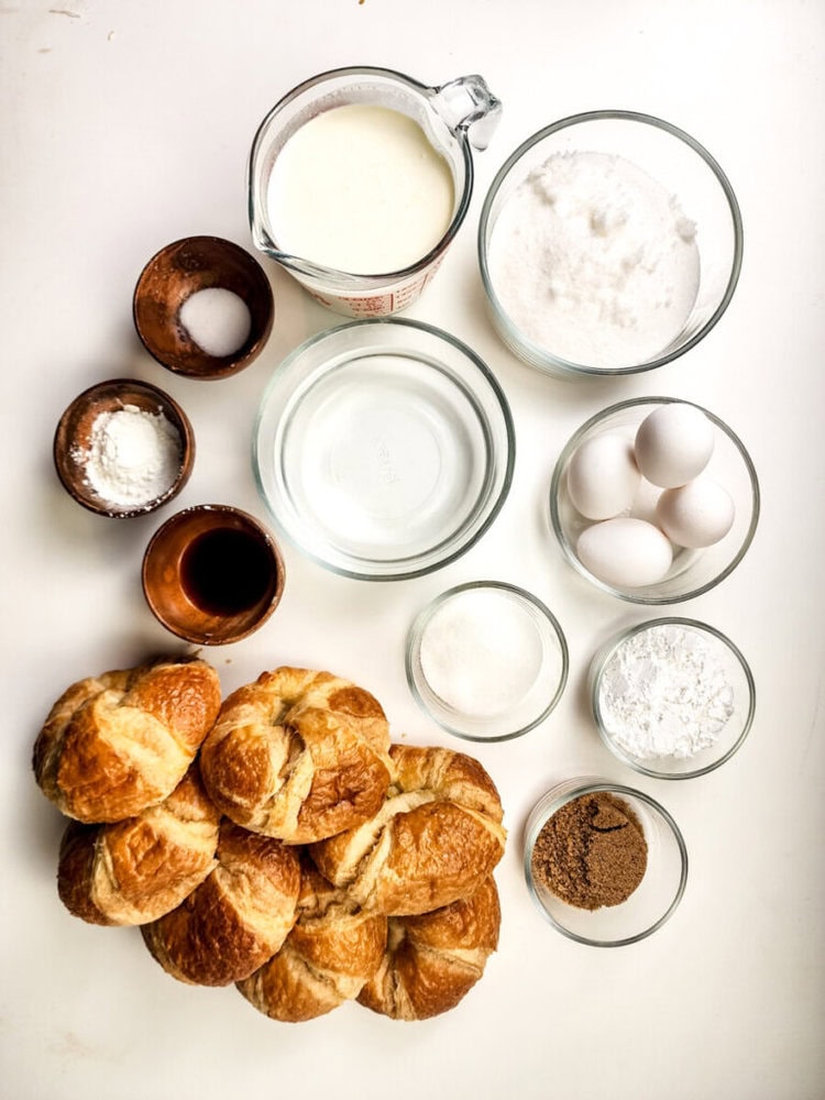 A top-down view of bowls and cups containing croissants, eggs, milk, sugar, vanilla, brown sugar, salt, and other baking ingredients arranged on a white surface.