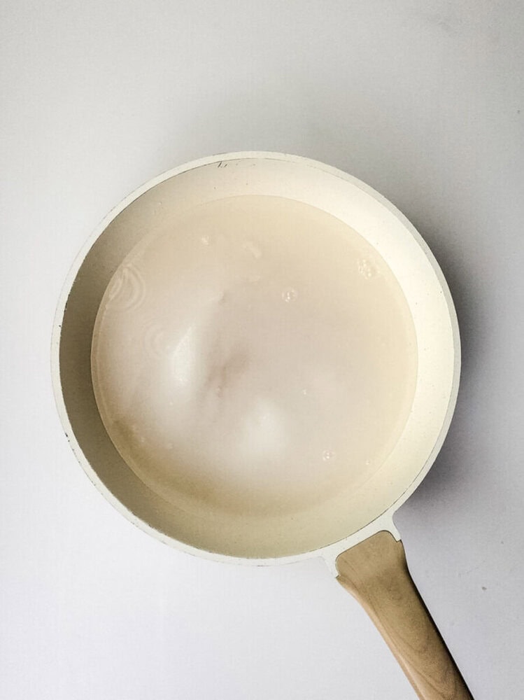 A white saucepan with a light wooden handle contains a cloudy, milky liquid on a plain white surface.