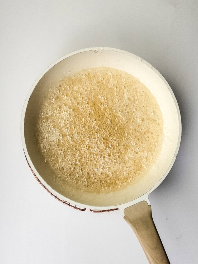 A white frying pan with a light brown, foamy liquid cooking inside, viewed from above on a white background.