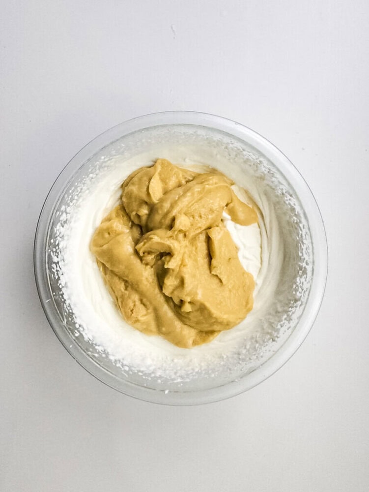 A glass bowl containing white whipped cream with a mound of light brown peanut butter mixture on top, set against a plain white background.