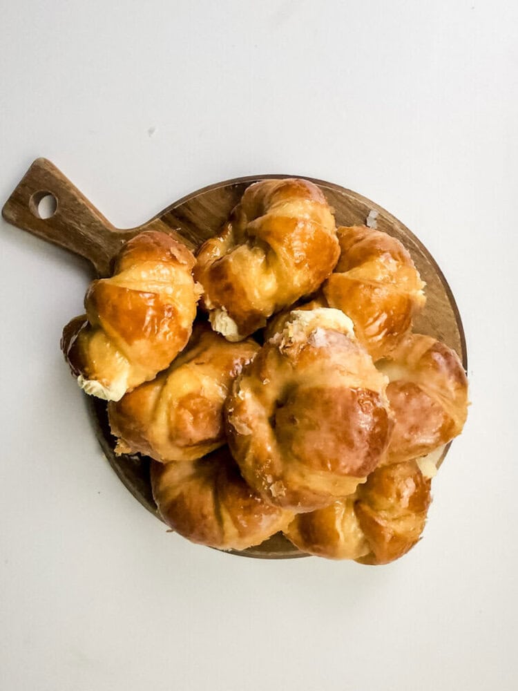 A wooden board holds a pile of golden-brown, glazed pastries with a creamy filling, set against a white background.