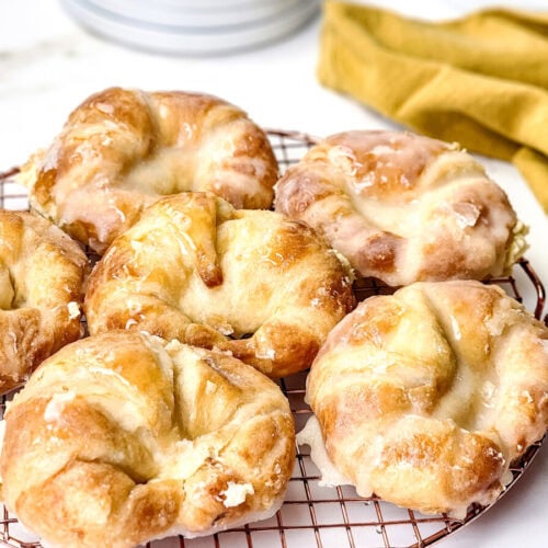 A batch of six glazed pastries cools on a round wire rack, with a yellow cloth and stacked plates in the background.