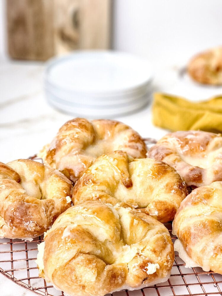 A close-up of several glazed pastries cooling on a wire rack, with stacked white plates and a yellow cloth in the blurred background.