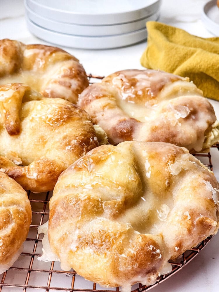 Four glazed pastries with a golden-brown crust rest on a cooling rack, with plates and a yellow cloth visible in the background.