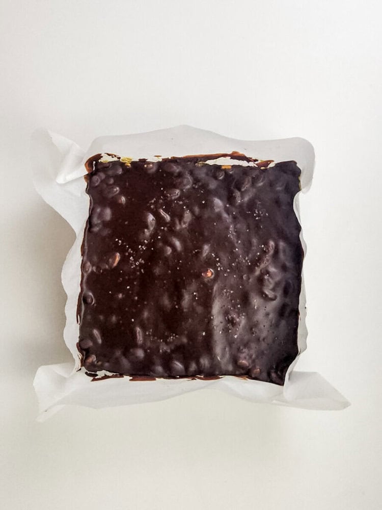 A square chocolate-covered dessert rests in a parchment-lined dish on a white background.