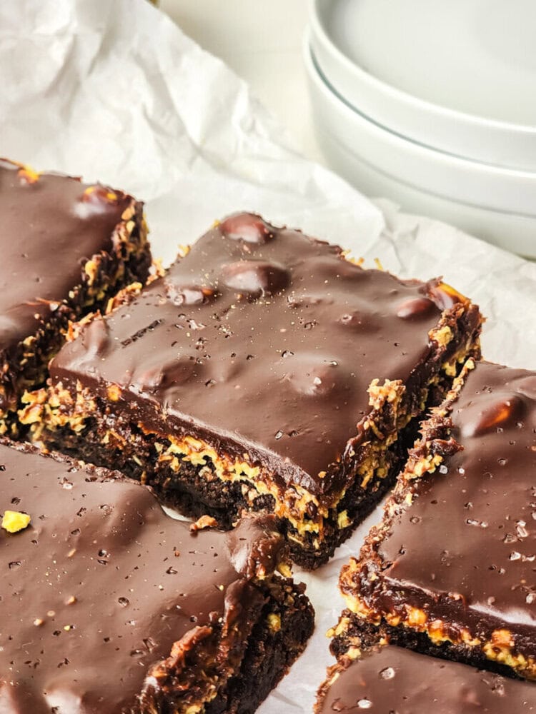Close-up of chocolate-covered brownies with a layered filling, cut into squares and placed on white parchment paper beside stacked white plates.
