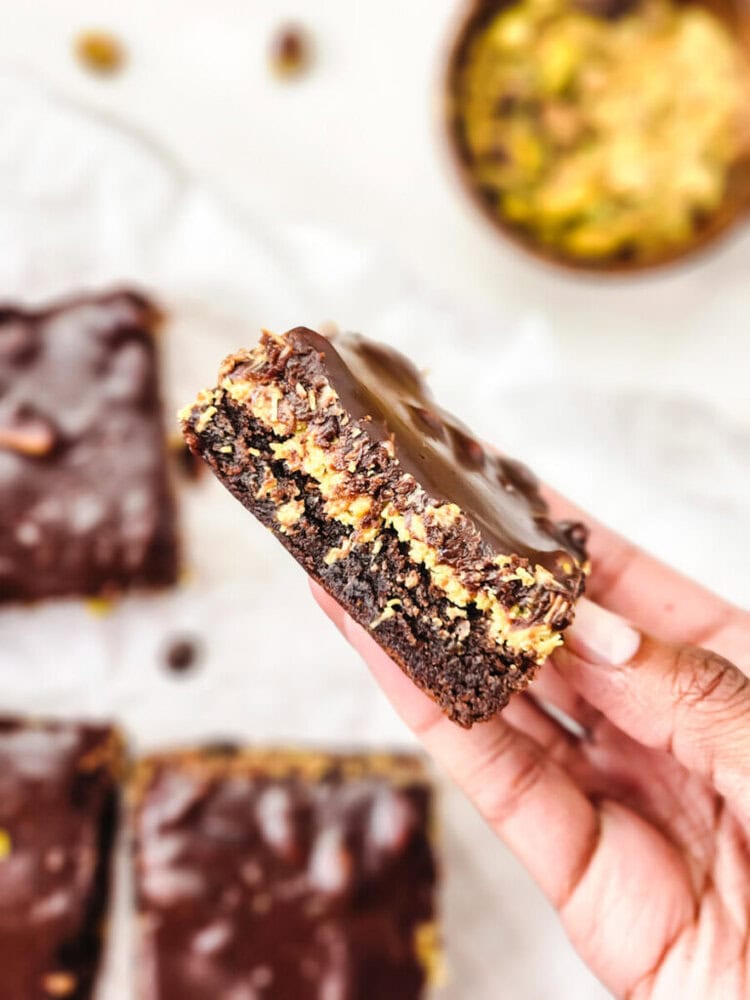 A hand holds up a chocolate dessert bar with visible layers of chocolate, a creamy filling, and a chocolate topping. More bars and a bowl are blurred in the background.