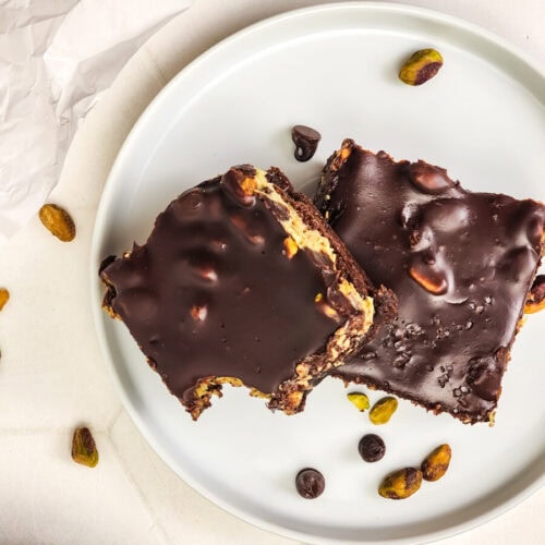 Two chocolate-covered dessert bars with visible nuts sit on a white plate, with scattered pistachios and chocolate chips on a white surface nearby.