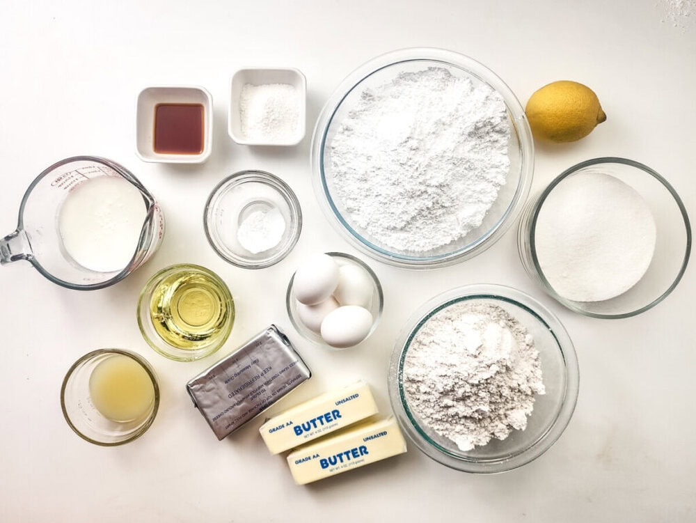 Various baking ingredients, including flour, sugar, eggs, butter, oil, milk, vanilla extract, cream cheese, lemon, and baking powder, arranged on a white surface.