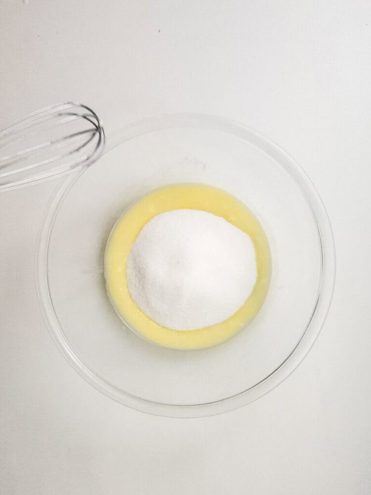 A glass bowl containing melted butter and a mound of white sugar sits on a white surface next to a metal whisk.