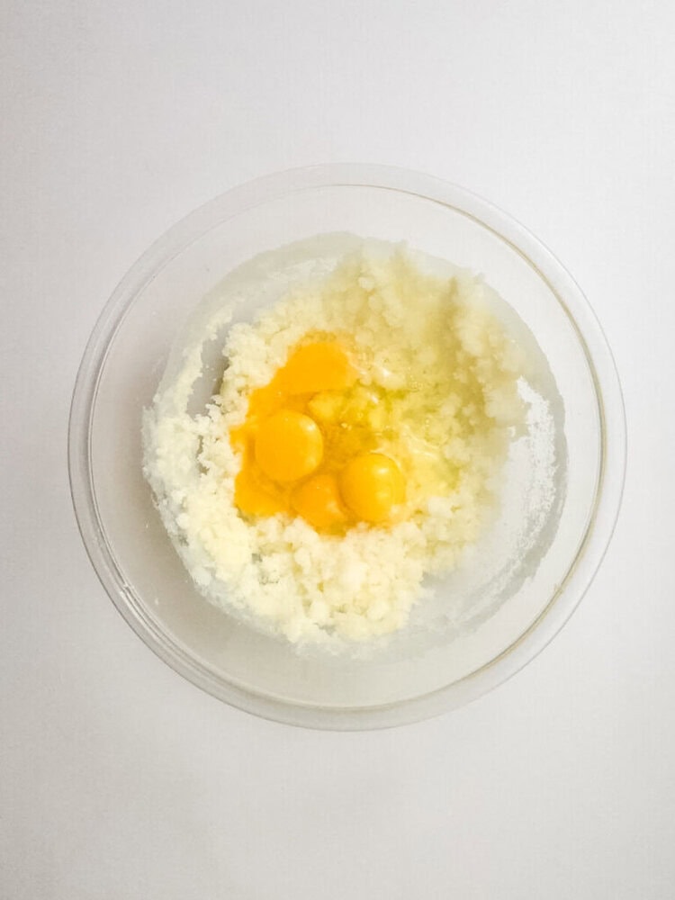 A clear bowl containing three raw eggs on top of a mixture of creamed butter and sugar, viewed from above on a white surface.