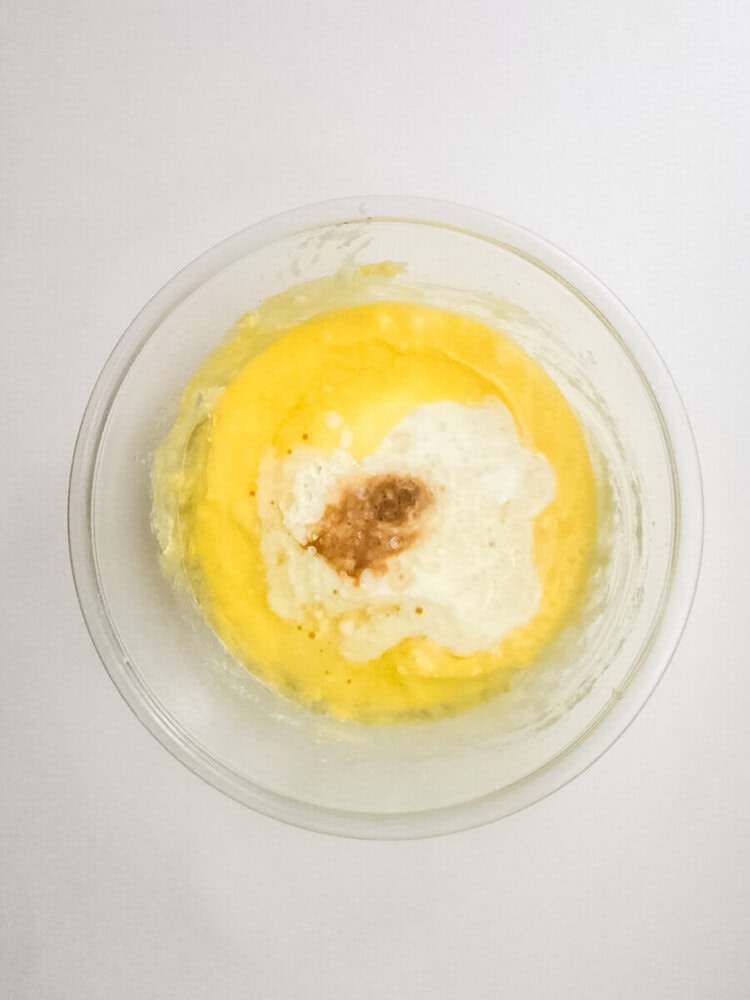 A glass bowl containing a mixture of beaten eggs, vanilla extract, and melted butter on a white surface.