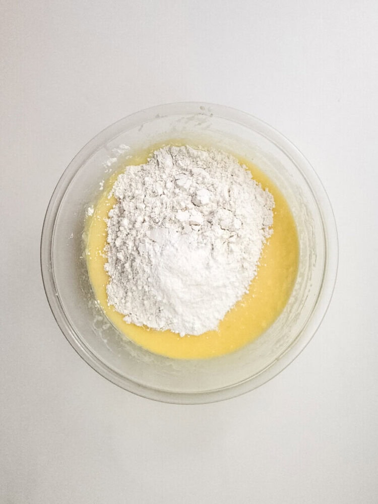 A clear glass bowl containing yellow batter with a mound of white flour on top, placed on a white surface.