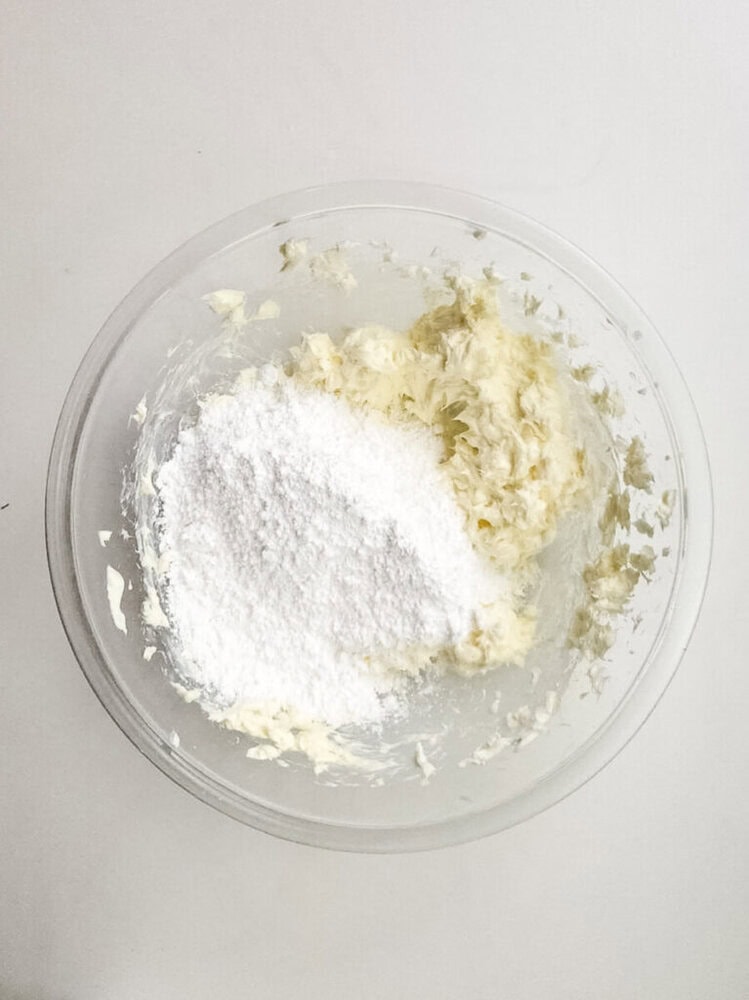 A glass bowl containing a mixture of softened butter and powdered sugar on a white surface.
