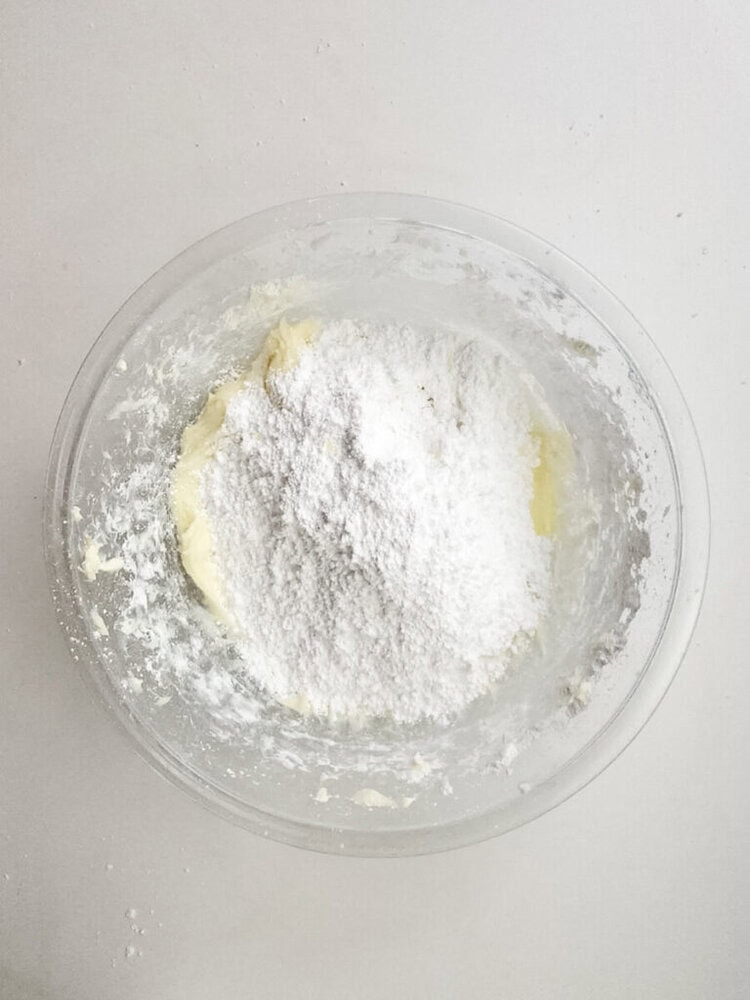 A glass bowl containing a mixture of butter and powdered sugar, sitting on a white surface.