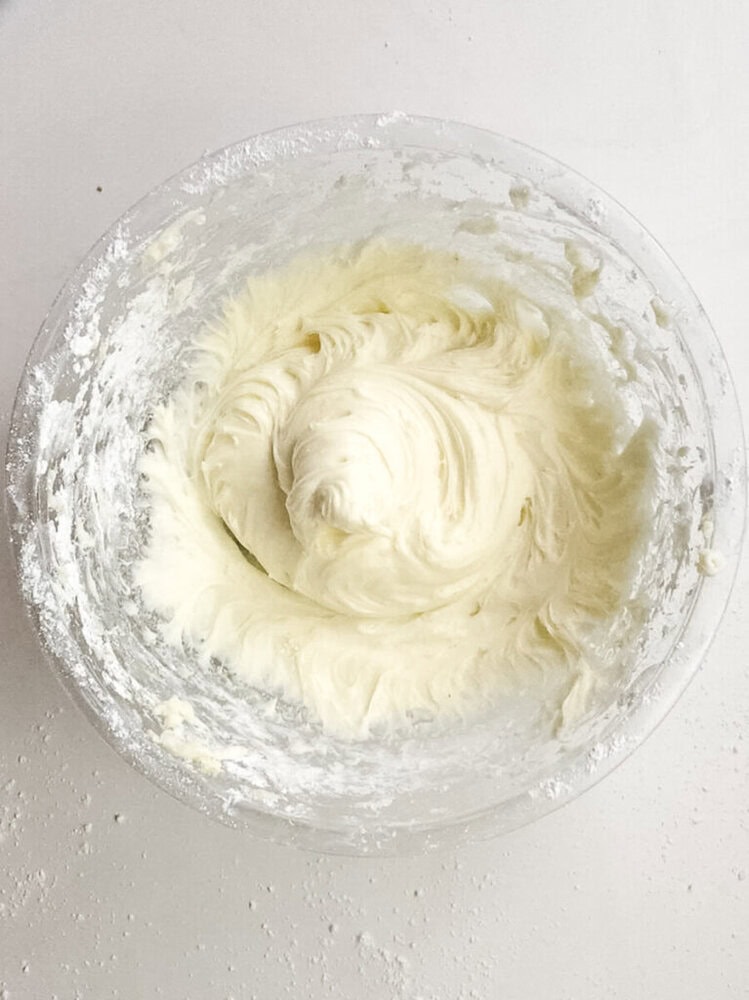 A glass mixing bowl containing whipped, creamy white frosting on a white surface, with some powdered sugar scattered around the bowl.