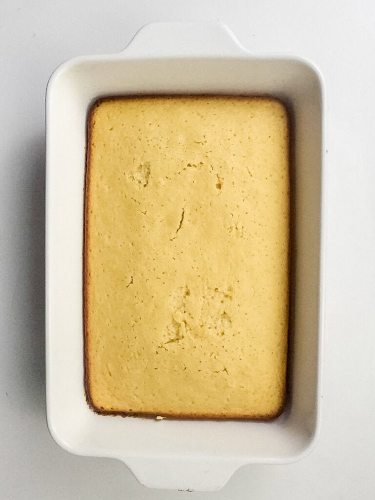 A rectangular cornbread loaf with a lightly browned edge sits in a white baking dish on a white surface.