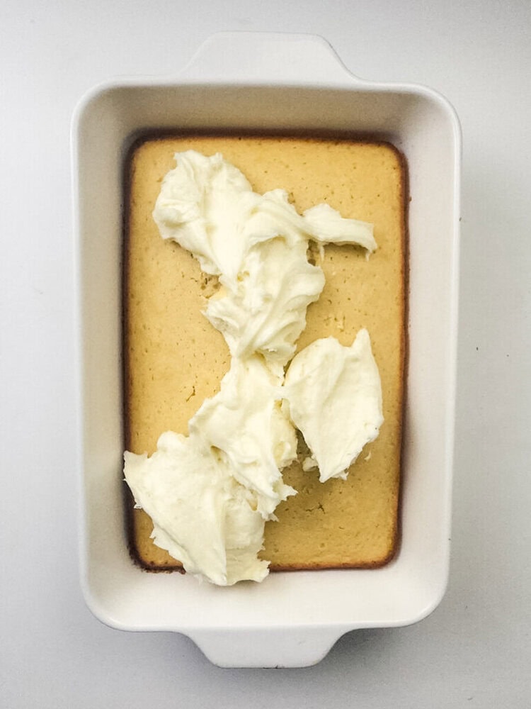 A rectangular yellow cake in a white baking dish with dollops of white frosting on top, not yet spread.