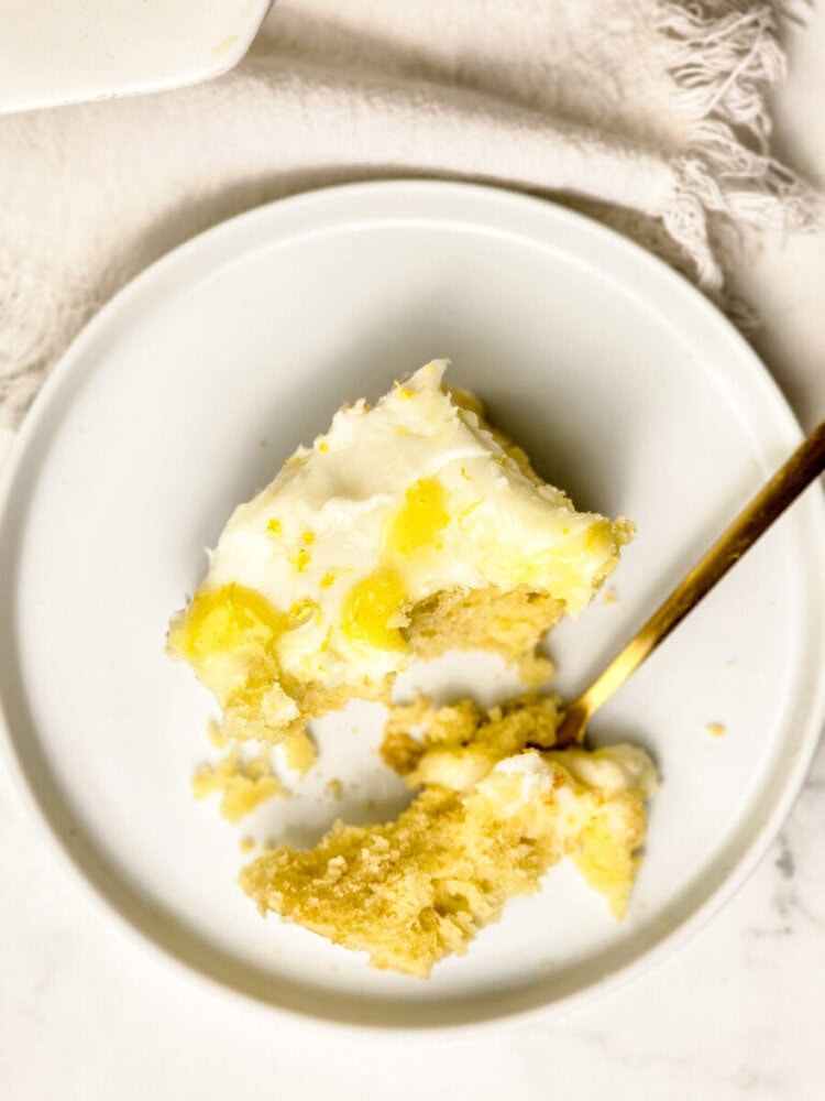 A slice of frosted yellow cake with a forkful taken out on a white plate, set on a white surface with a fringed cloth nearby.