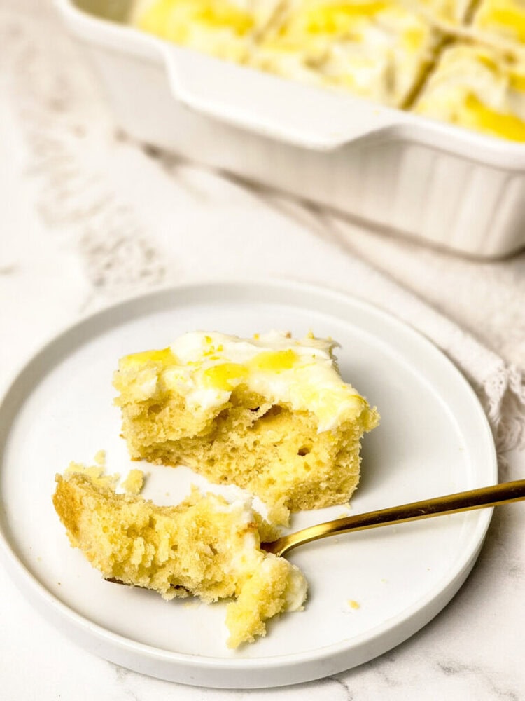A piece of lemon-flavored cake with white icing sits on a white plate with a gold fork. The cake pan is visible in the background.