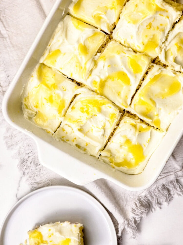 A white baking dish with frosted lemon bars cut into squares, some with visible swirls of yellow, sits on a textured white cloth next to a plate holding one piece.