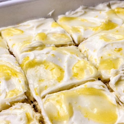 A close-up view of lemon bars topped with creamy white frosting and yellow lemon zest, cut into square pieces in a baking dish.