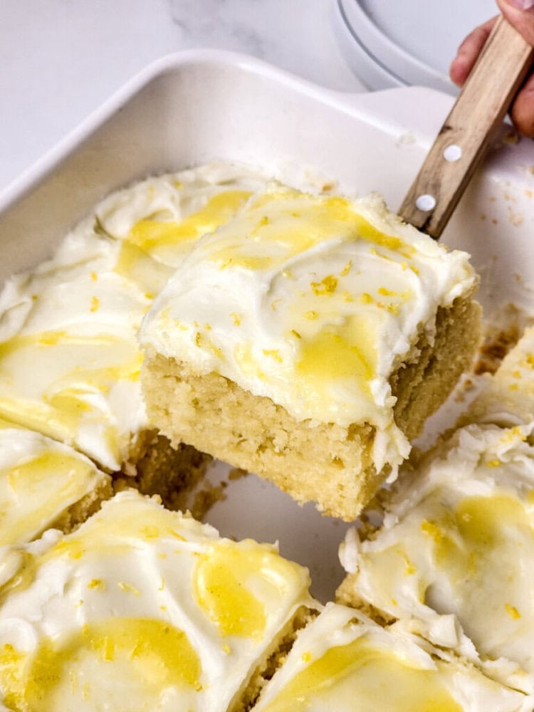 A hand lifts a square piece of frosted lemon cake from a baking dish, showing a moist, light crumb and swirls of yellow lemon zest on top.