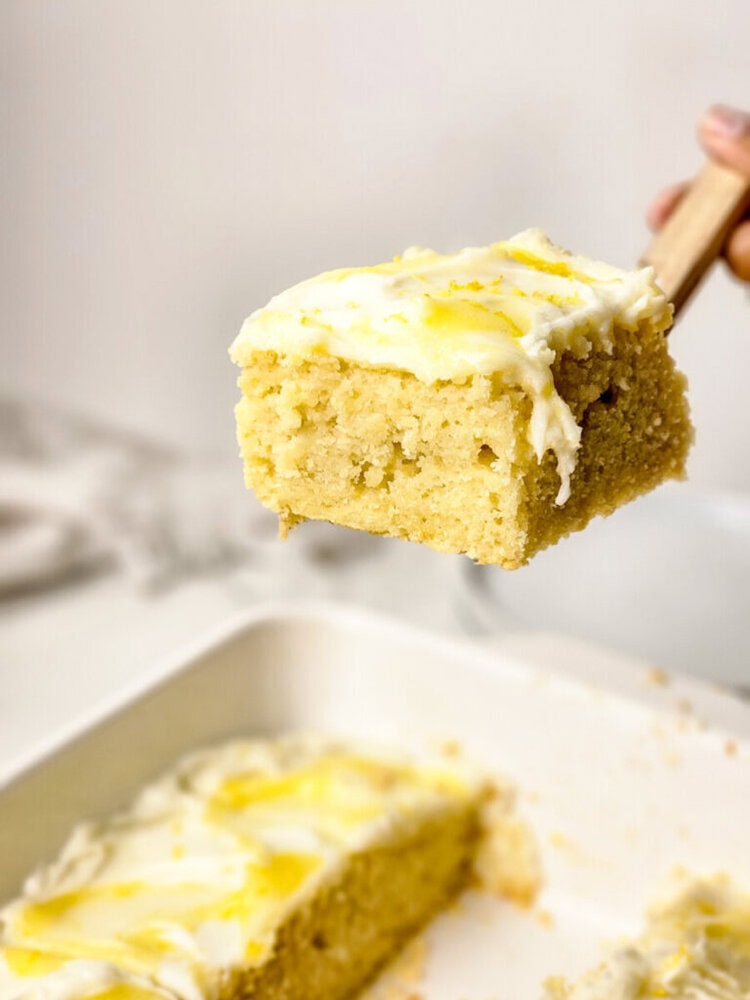 A square piece of yellow cake with white frosting and lemon zest on a spatula, held above a baking dish with more cake inside.