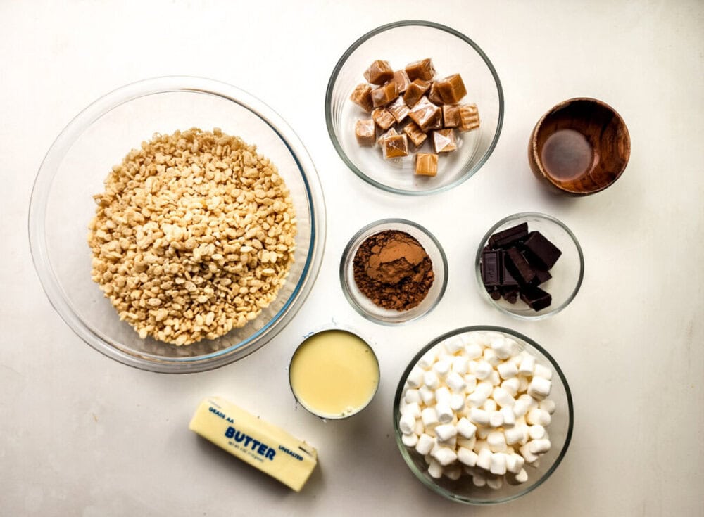 A variety of ingredients in bowls, including crispy rice cereal, caramels, cocoa powder, chocolate, mini marshmallows, sweetened condensed milk, butter, and vanilla extract.