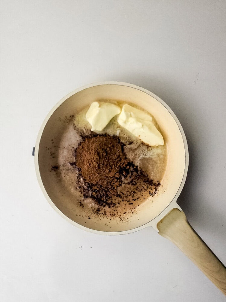A white pan contains butter, cocoa powder, sugar, and a small amount of flour or powder on a white surface, ready to be mixed.