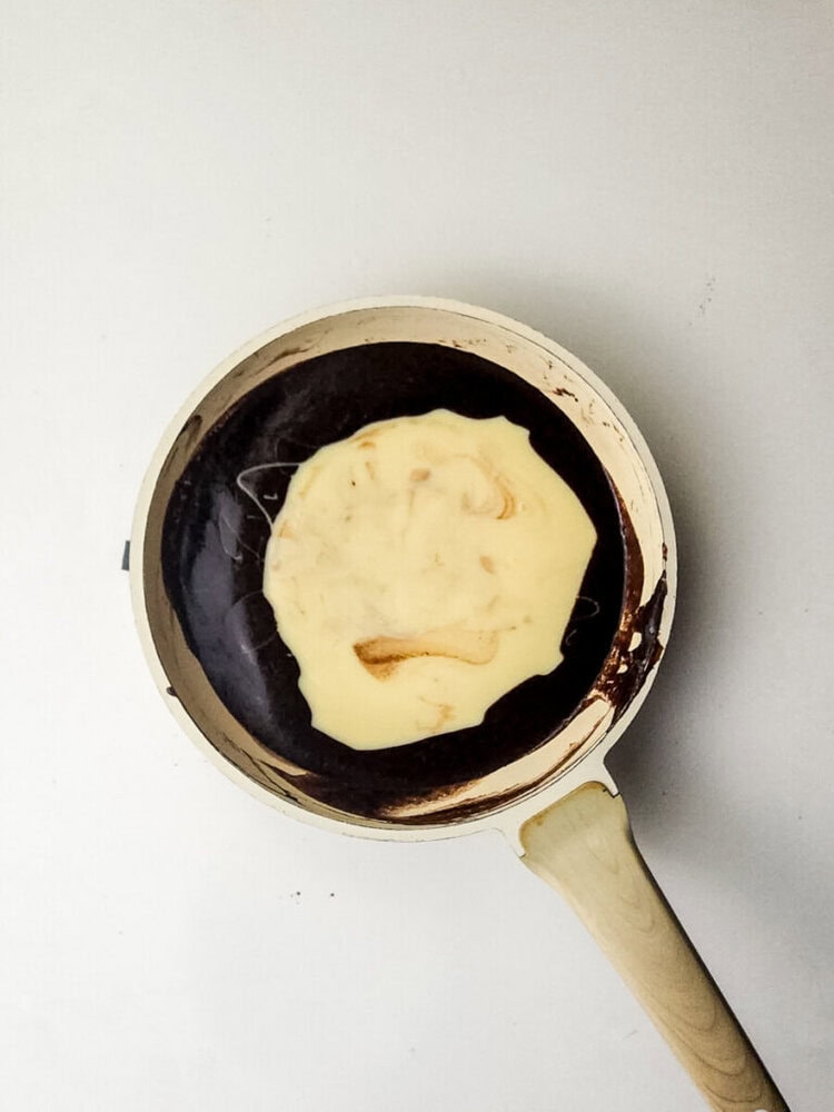 A pancake cooking in a light-colored frying pan on a white surface.