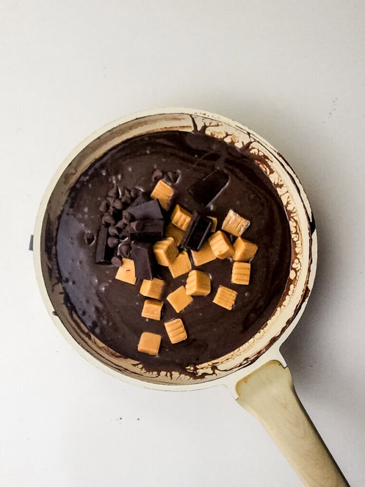 A pan filled with chocolate batter, topped with chocolate chunks, chocolate chips, and caramel candies, on a white surface.