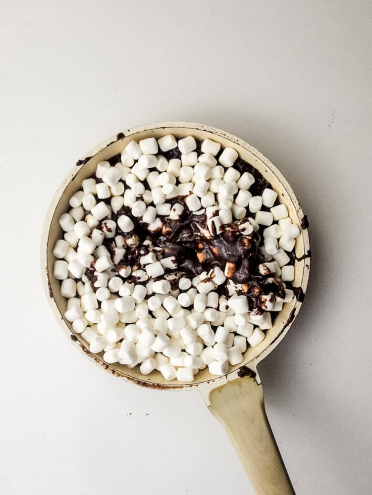 A pan filled with melted chocolate and topped with mini marshmallows sits on a white surface.