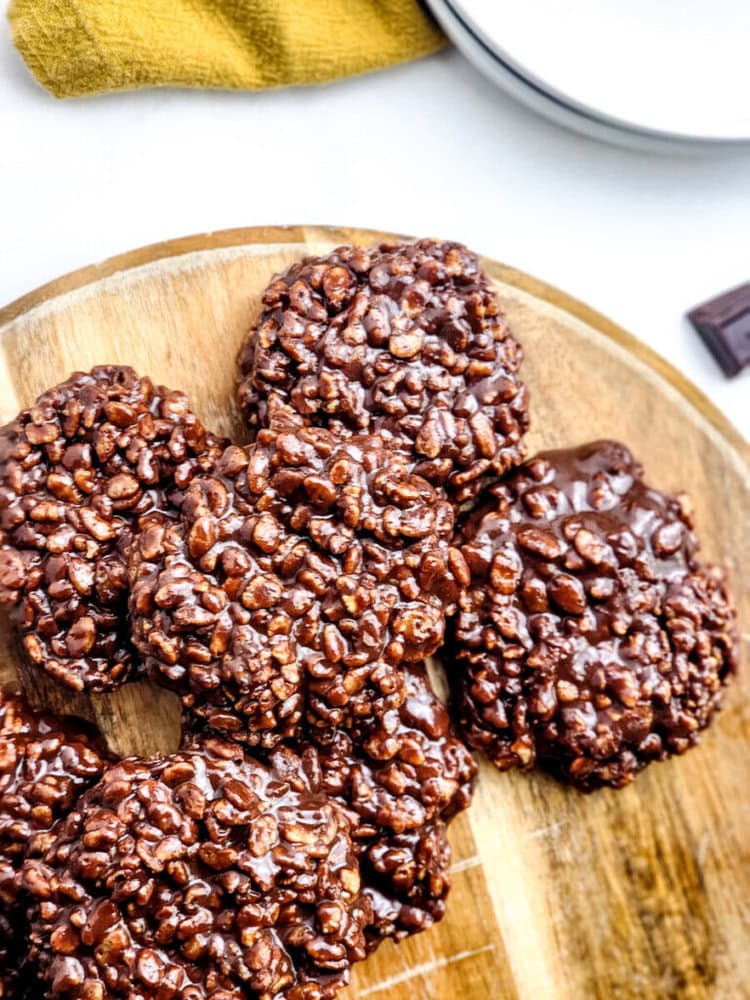 Chocolate rice crispy clusters are arranged on a wooden board, with a yellow napkin and white plates in the background.