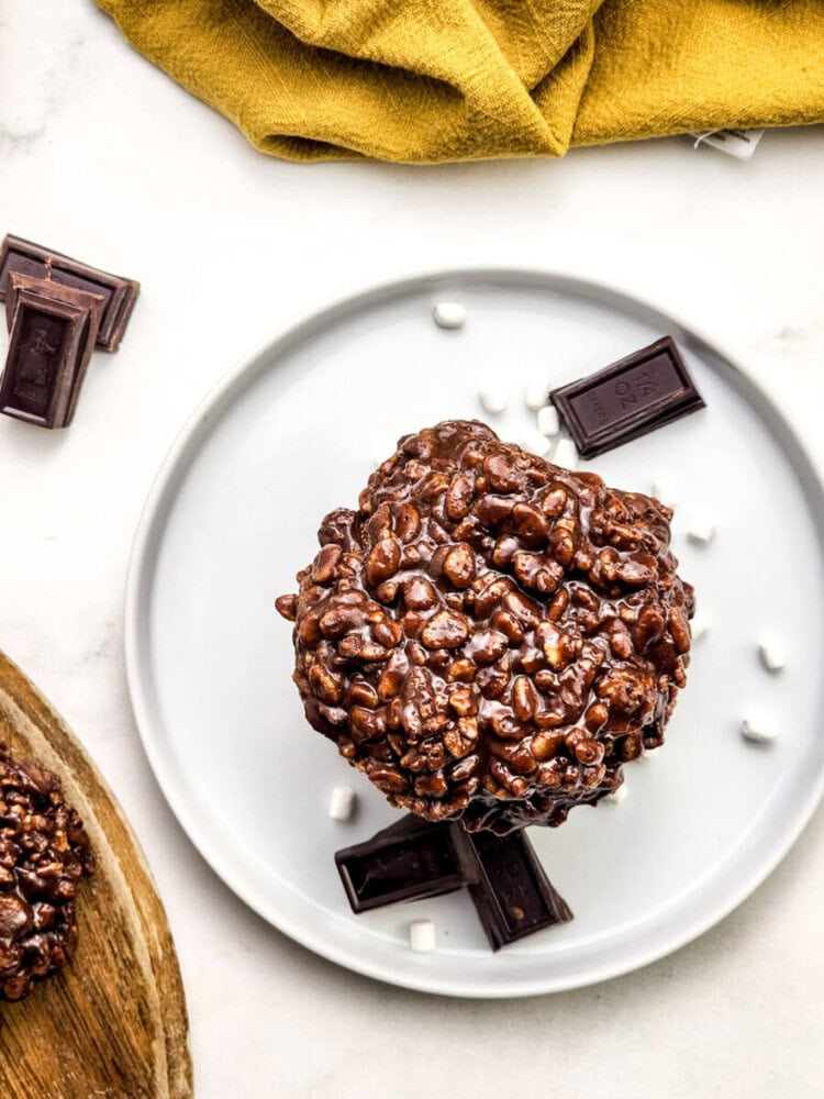 A large chocolate rice crispy treat sits on a white plate with chocolate pieces and mini marshmallows, next to a yellow cloth.