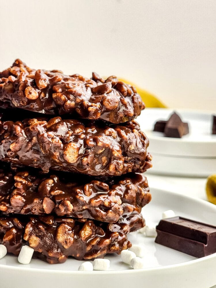 A close-up of a stack of chocolate-covered oat and marshmallow cookies on a white plate, with pieces of chocolate and mini marshmallows nearby.