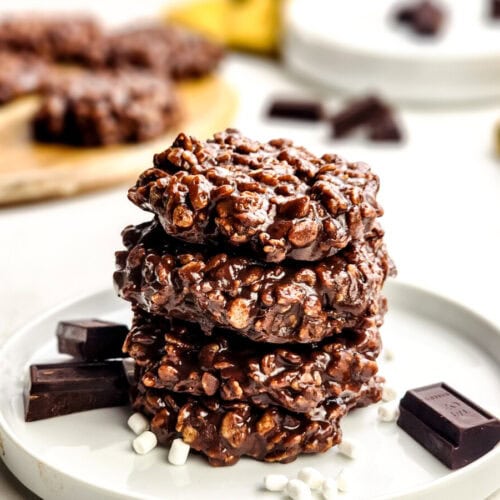 Three chocolate rice crispy cookies are stacked on a white plate, surrounded by chocolate pieces and a few mini marshmallows. More cookies and chocolate are in the blurred background.