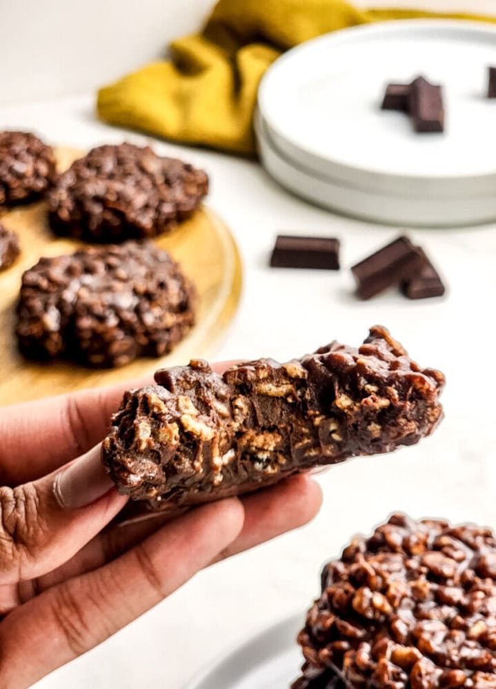A hand holds a no-bake chocolate oatmeal cookie, with more cookies and chocolate pieces in the background on a white surface.